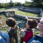 2 jeunes garçons et leurs 2 mamans découvrant les vestiges d'une maison romaine in situ, à l'aide d'une application de réalité virtuelle sur tablette.
